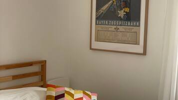 A bed with white bedding, a wooden headboard, and colorful pillows. A framed poster of a mountain landscape is on the wall.