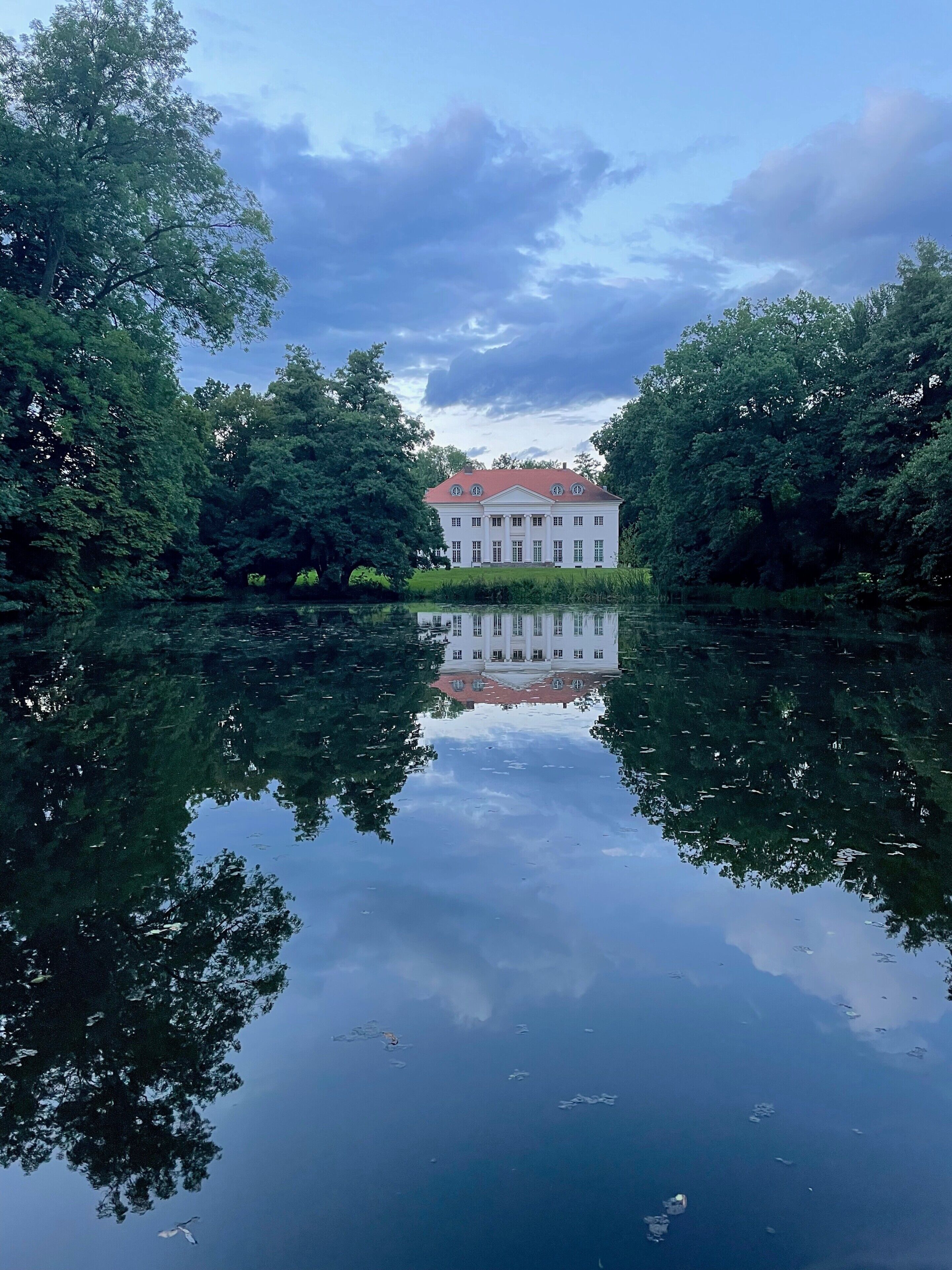 A large, symmetrical building with a red roof, surrounded by dense trees and reflecting in calm water.