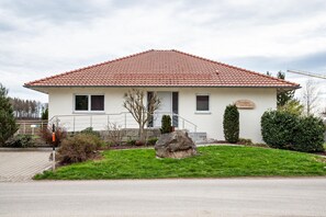 Une maison de plain-pied avec un toit recouvert de tuiles rouges, une porte d’entrée et un petit jardin agrémenté de rochers et d’arbustes.