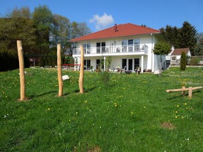 Une maison à deux étages avec un toit rouge, un balcon et un jardin où poussent des fleurs jaunes.