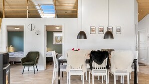 A spacious room with a wooden ceiling, a dining area with white chairs and a fur throw, a bed in the adjacent room, and a skylight.