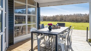 A covered outdoor patio with a table and chairs, a large sliding glass door, and a view of a grassy field.