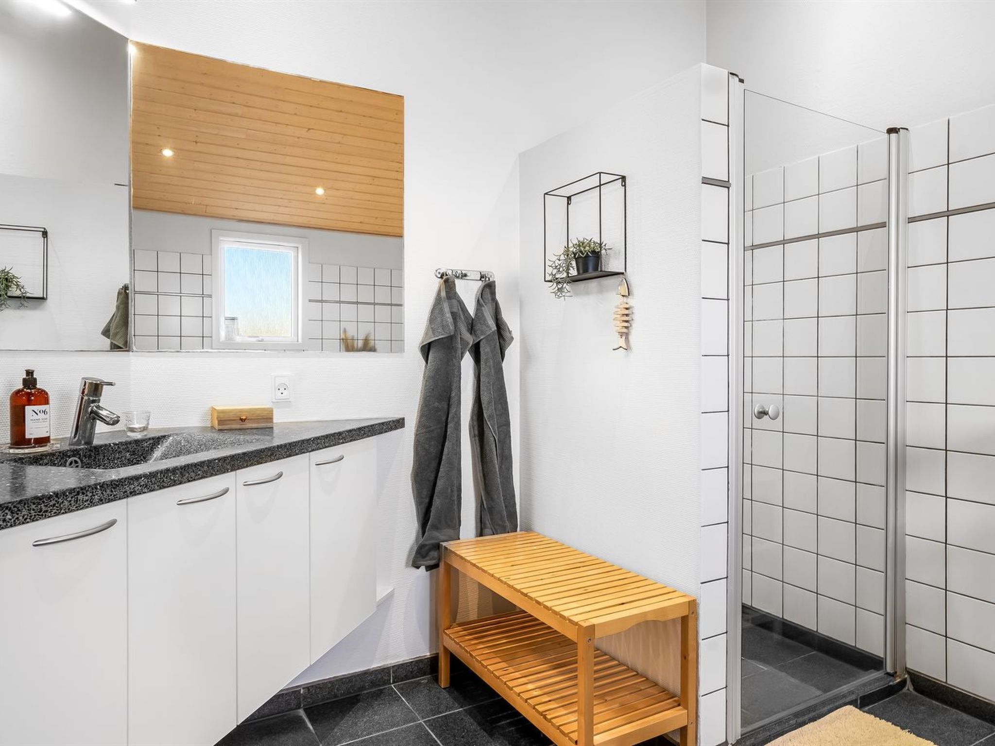 Une salle de bain moderne avec un comptoir en granit, des armoires blanches, un banc en bois, une cabine de douche en verre et une petite étagère avec une plante.