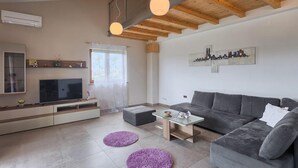 A modern living room with a grey sofa, a TV, and a coffee table. There is a window with sheer curtains and a cityscape painting on the wall.