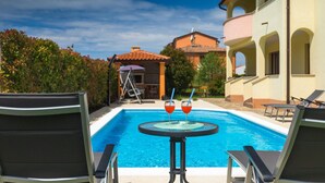 A poolside view with two lounge chairs, a table with two glasses of red drink, and a building with a tiled roof in the background.