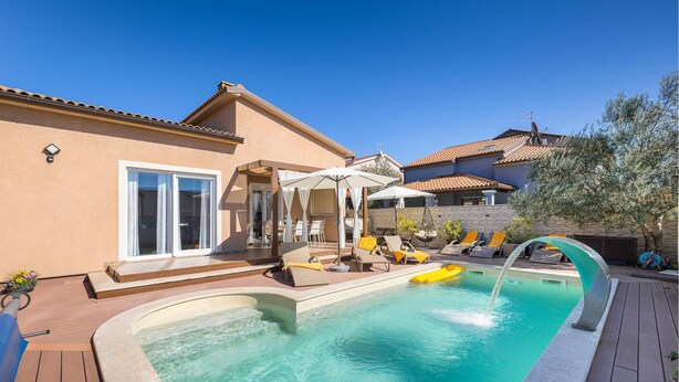Une piscine avec des chaises longues et des parasols, un espace patio et une maison avec un toit en tuiles.