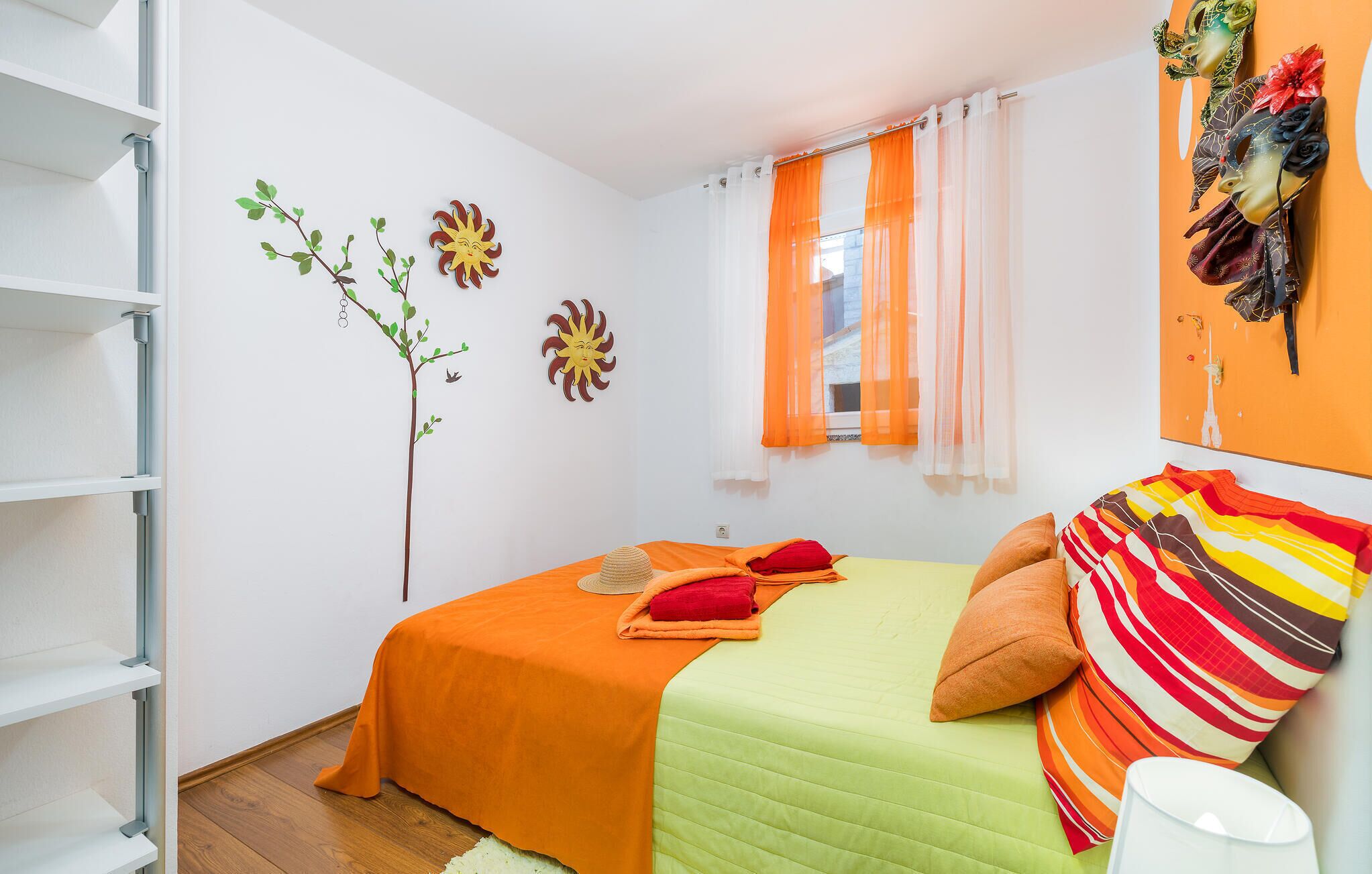 A bedroom with a bed, a window with orange curtains, a wall with a sun decoration, and a shelf unit.