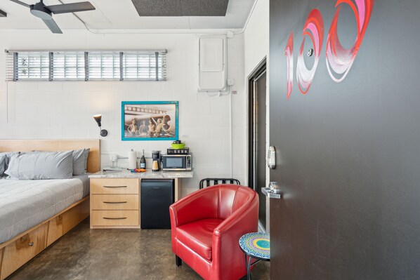 Habitación de hotel pequeña con cama, escritorio, una silla roja, una mesita y una puerta con el símbolo de número.