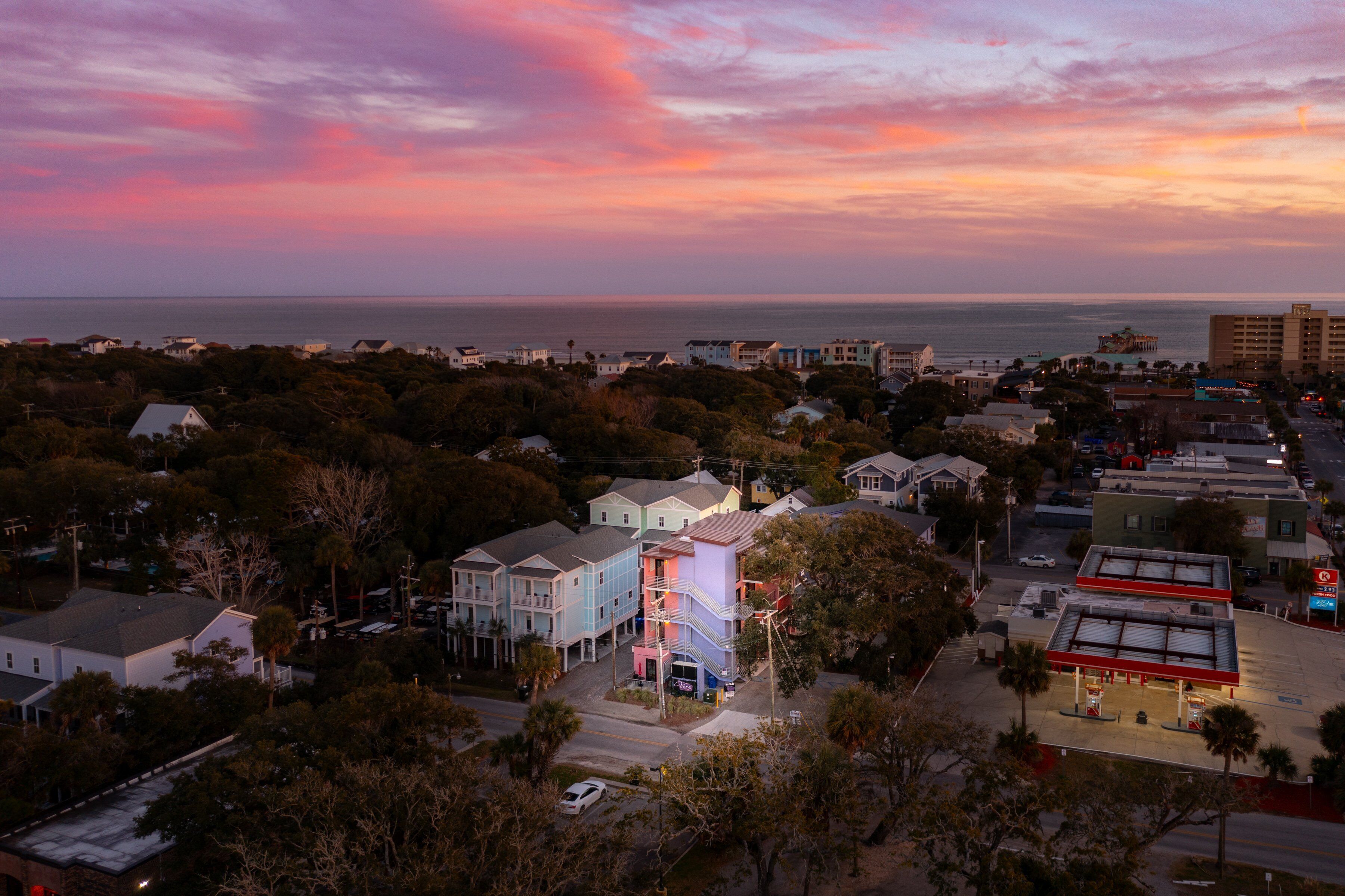 An aerial view of a coastal town with residential buildings, a gas station, and a view of the ocean during sunset.