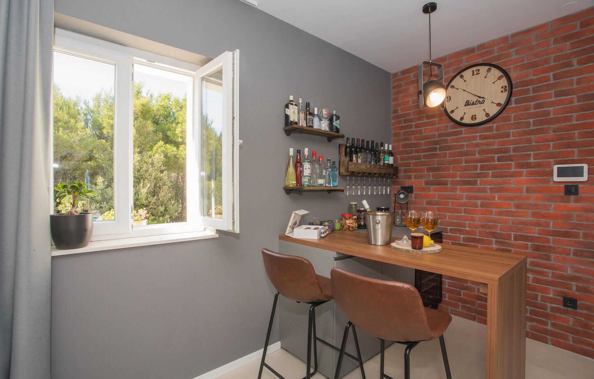 Cocina con barra de madera, dos taburetes, un reloj y ventana con vistas a follaje.