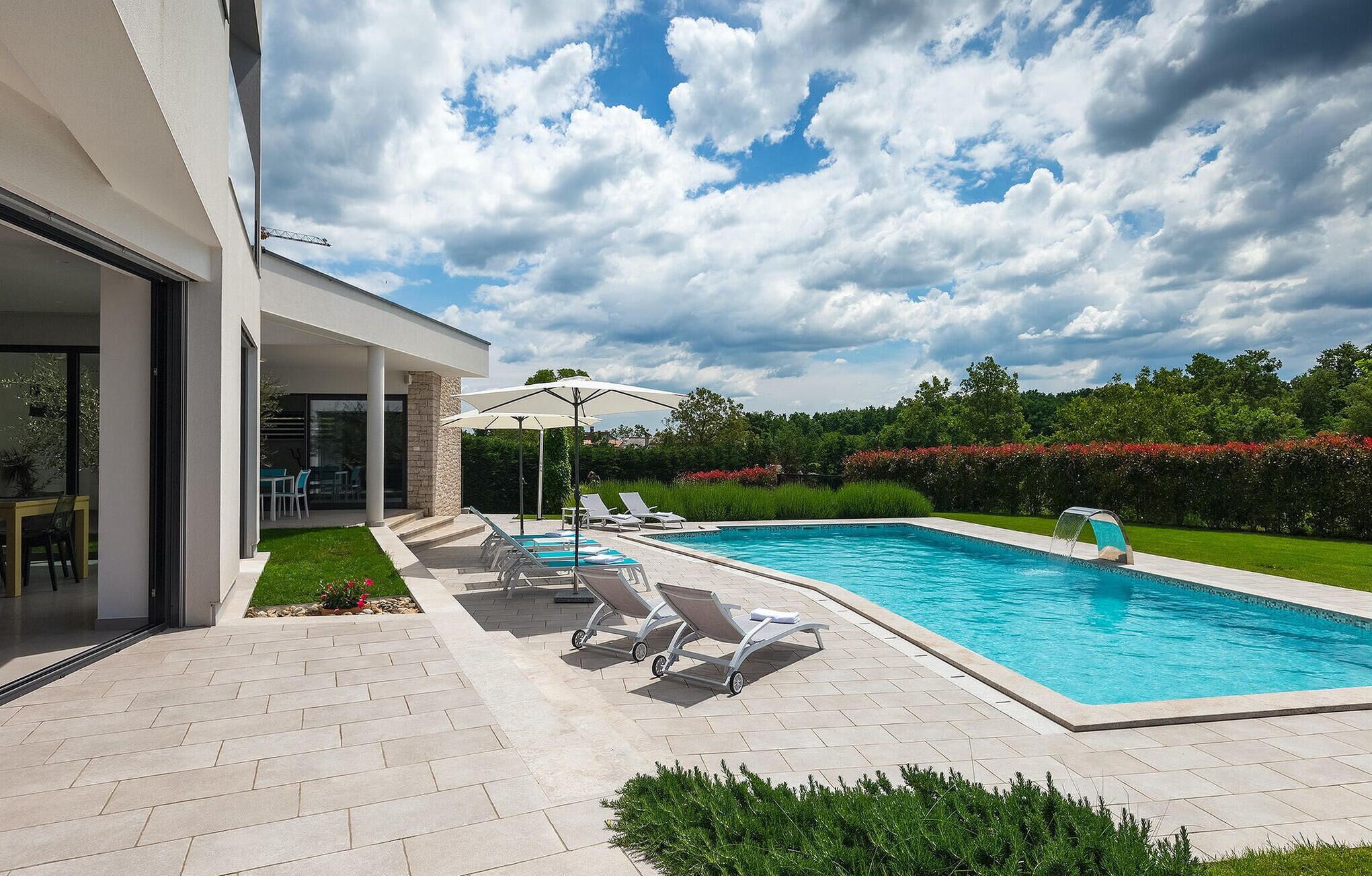A modern outdoor pool area with lounge chairs and umbrellas, surrounded by a well-maintained lawn and hedges.