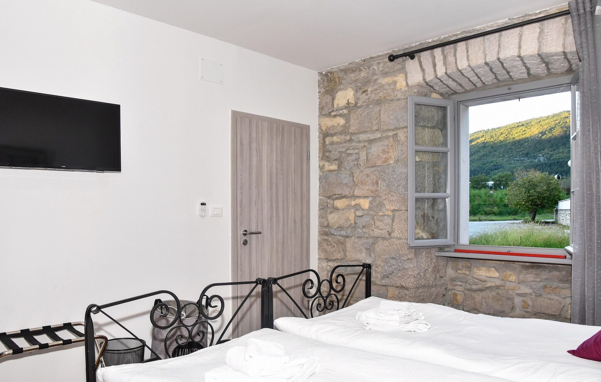 A bedroom with a bed, a TV mounted on the wall, a stone wall, and a window with a view of greenery.