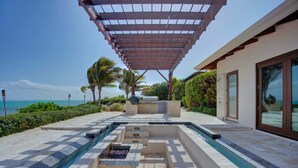 Zona de piscina con pérgola, vista al océano y un edificio moderno con amplios ventanales.