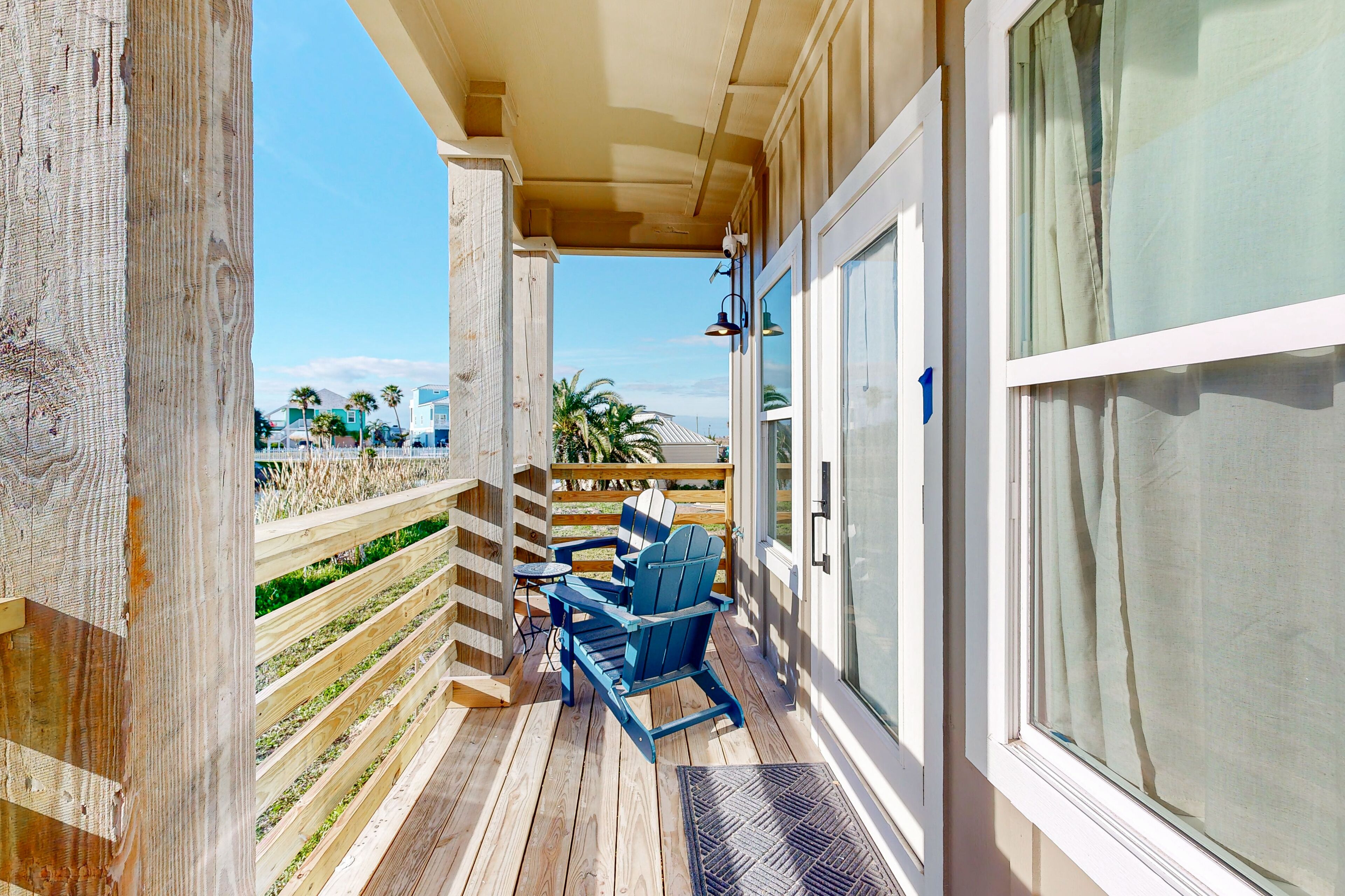 Um deck de madeira com uma cadeira Adirondack azul e uma pequena mesa, com vista para uma praia com palmeiras e casas.