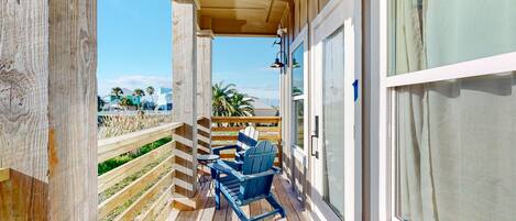 A wooden deck with a blue Adirondack chair and a small table, overlooking a beach with palm trees and houses.