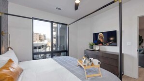A modern bedroom with a bed, a TV, a ceiling fan, and a view of the city.