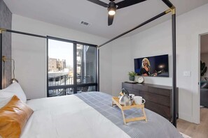 A modern bedroom with a bed, a TV, a ceiling fan, and a view of the city.