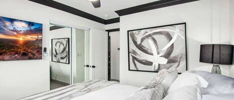 A modern hotel room with a bed, ceiling fan, artwork, and a door leading to another room.