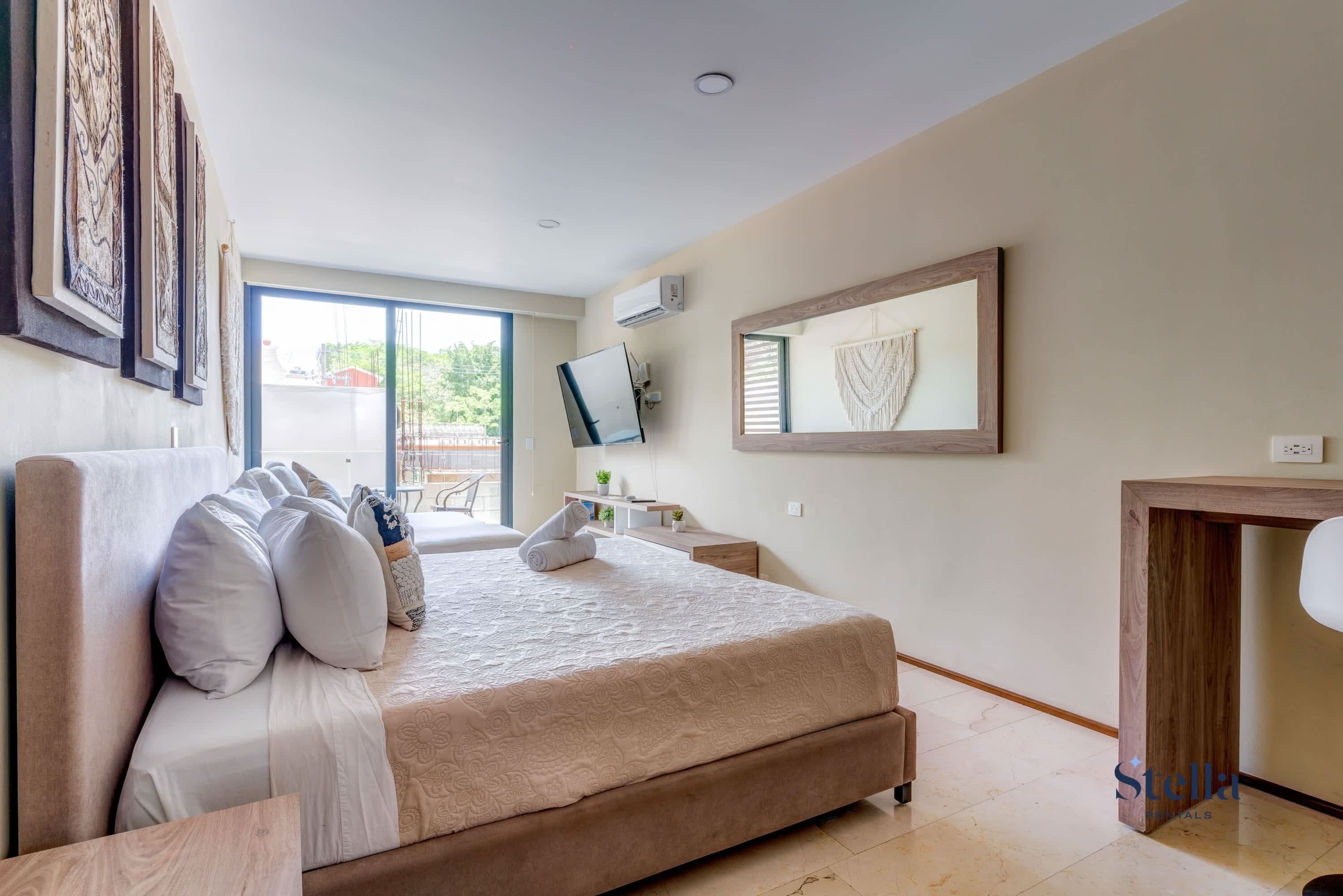 A modern bedroom with a large bed, a TV, a desk, and a view of the outdoors.
