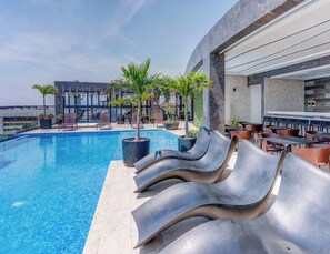 Área de piscina com espreguiçadeiras, espaço para refeições e um edifício de design moderno.