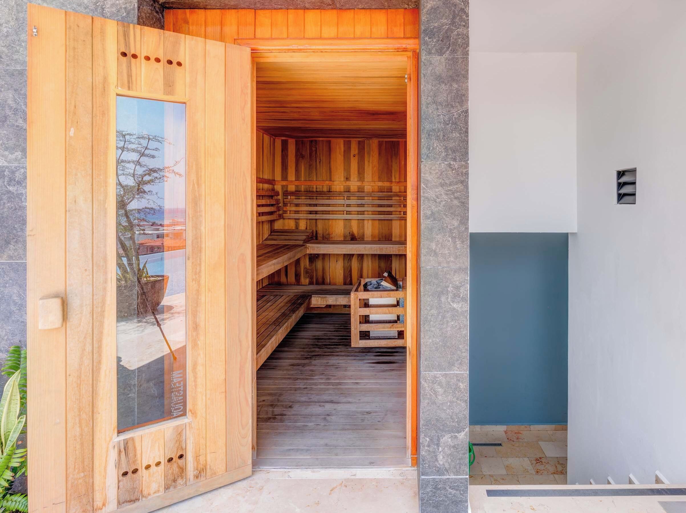 Una sauna de madera abierta con puerta de vidrio, pared de piedra y pared azul contigua.
