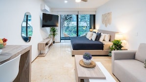 A modern hotel room with a bed, a sofa, a TV, a ceiling fan, and a large window offering a view of the outdoors.