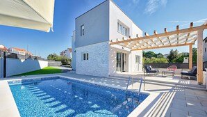 Un espace piscine extérieur moderne, doté d’une pergola, d’un espace salon couvert et d’un bâtiment blanc à la façade en pierre.