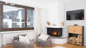 A modern living room with a fireplace, two grey bean bag chairs, a TV mounted on the wall, and a window with a view of buildings.