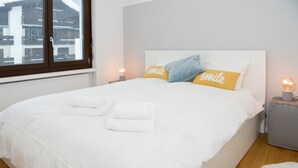 A neatly made bed with white bedding and two pillows, one yellow and one blue, in a room with a large window and a bedside lamp.