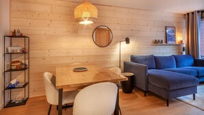 A cozy living room with a wooden wall, a round mirror, a blue sofa, a dining table with chairs, and a hanging light fixture.