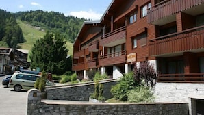 A multi-story wooden building with balconies, a stone wall in front, and a parking area with cars.