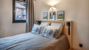 A bedroom with a bed, a window with curtains, a wall-mounted lamp, and three framed pictures.