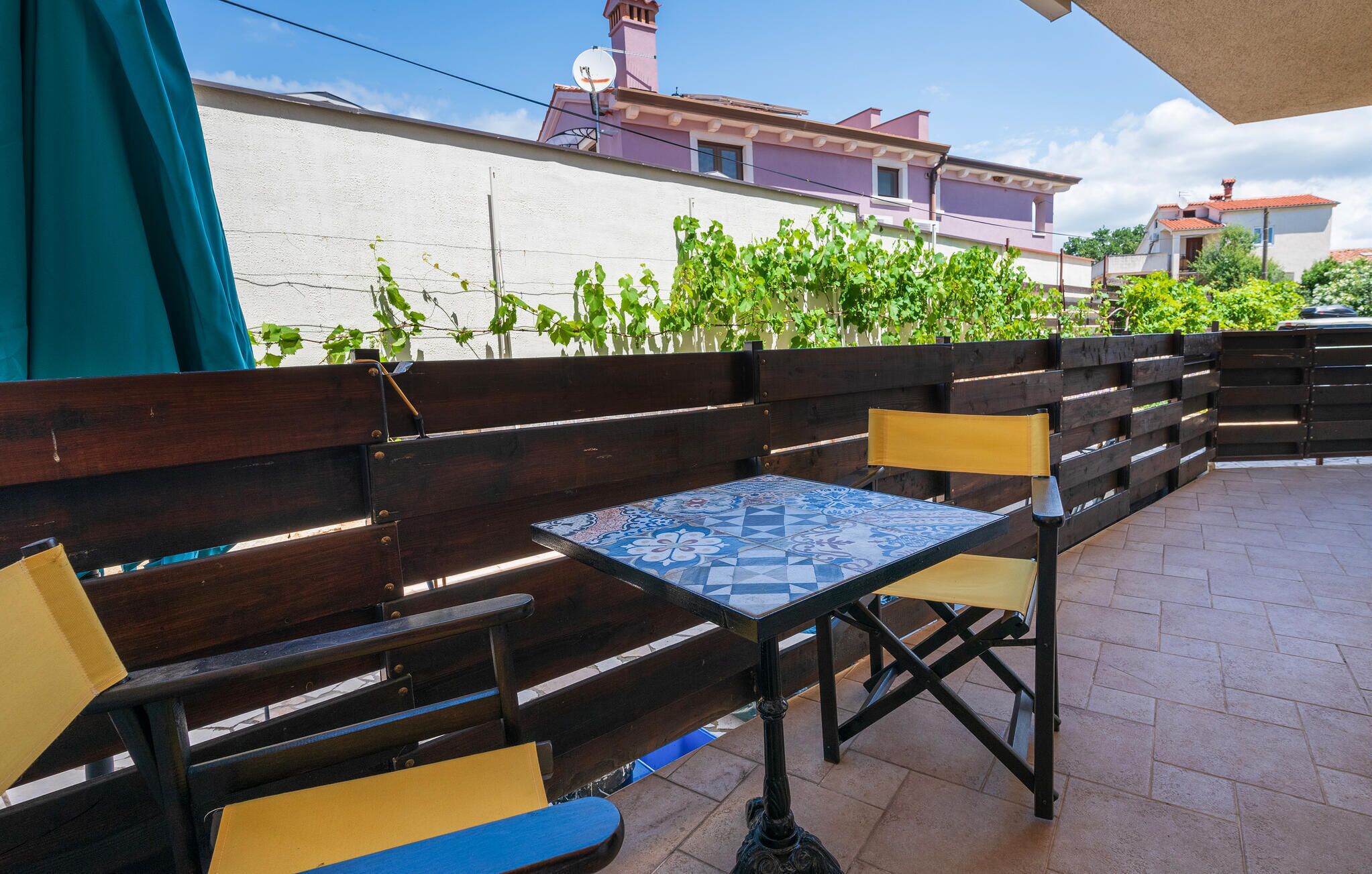 Un espace patio avec des bancs en bois et une table carrelée, entouré d’un petit muret et de verdure.