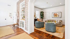 A spacious living area with a brick accent wall, a wooden floor, a seating area with a sofa and ottoman, and a kitchen visible through an archway. - 6A at Saint Philip Square, One Block to King, Classic Charleston Comfort (Charleston)