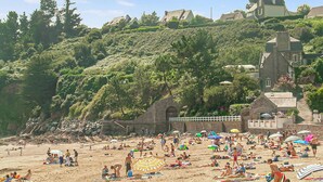 Ein Strand mit badenden und sonnenden Menschen, umgeben von üppiger Vegetation und einem Hügel mit Gebäuden.