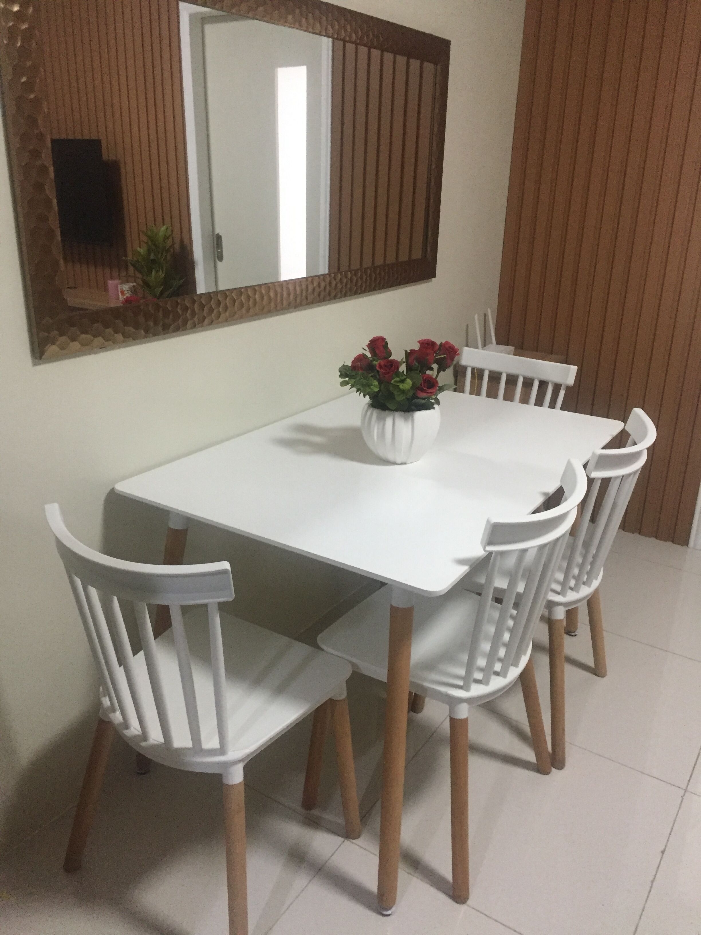 A dining area with a white table and chairs, a vase with red flowers, and a mirror on the wall.