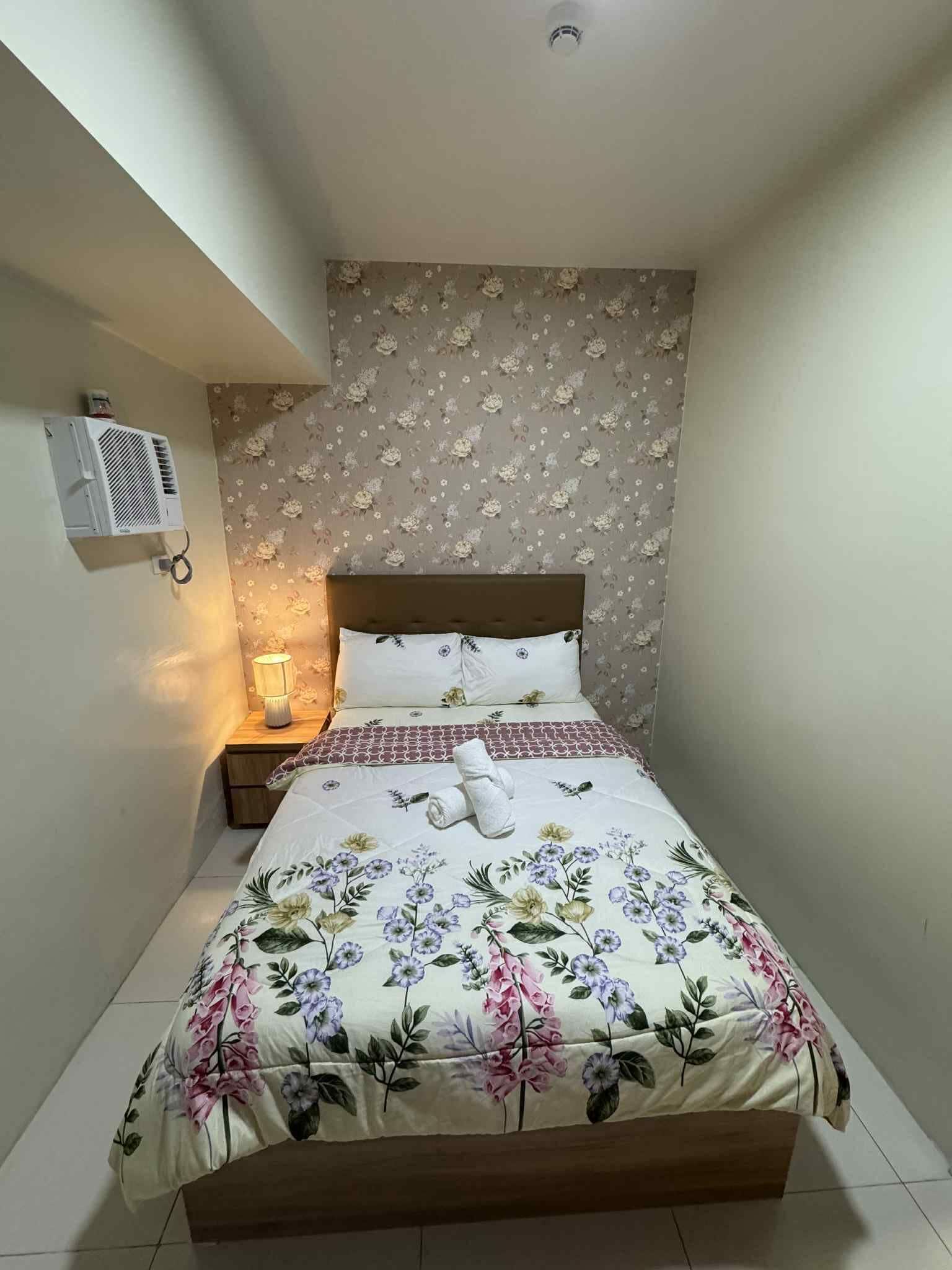 Une petite chambre à coucher, bien rangée, avec une couvre-lit à motifs floraux, une lampe de chevet et un climatiseur fixé au mur.