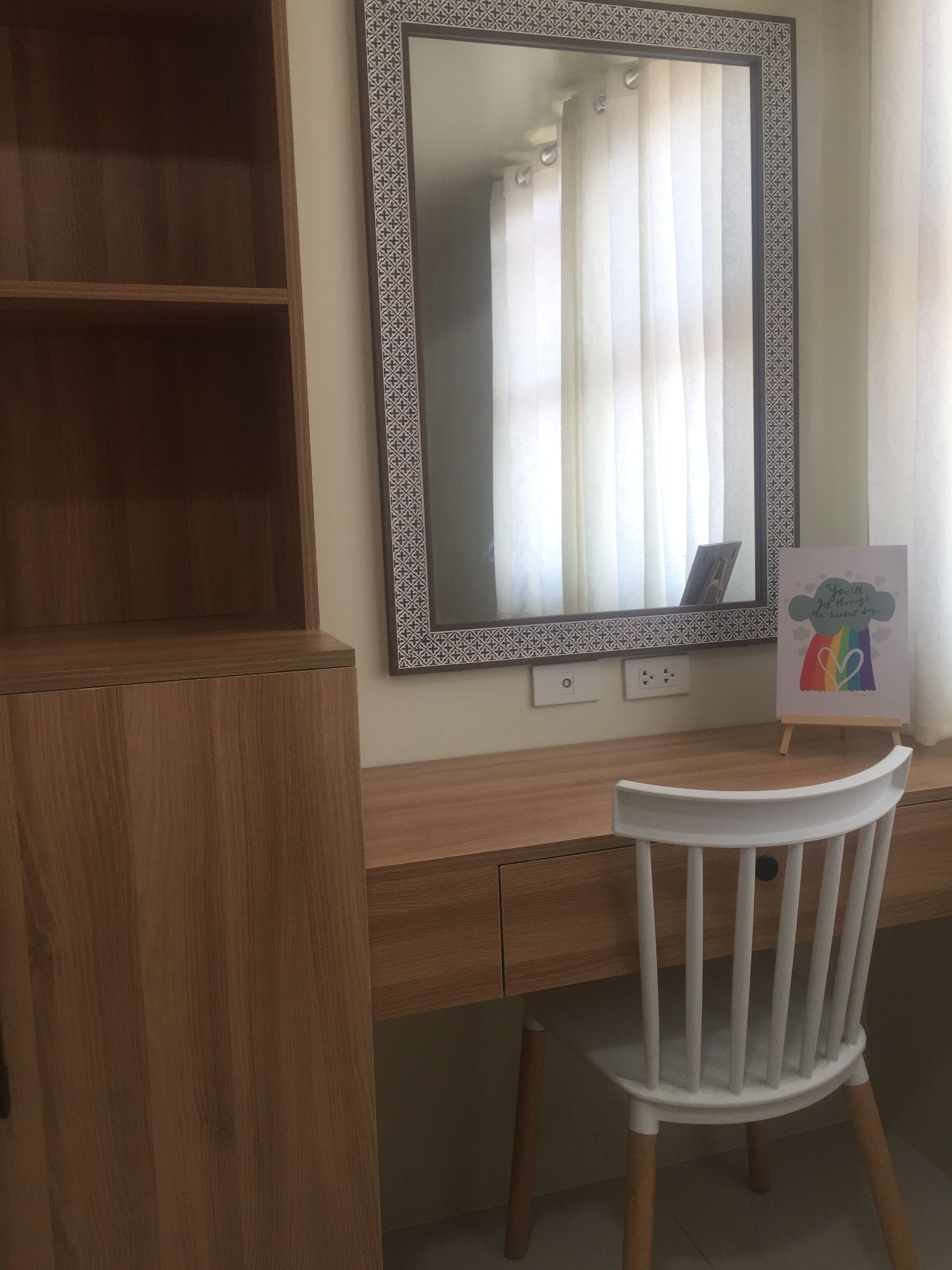 A wooden desk with a white chair, a large mirror with a patterned frame, and a small easel with a rainbow and heart drawing.