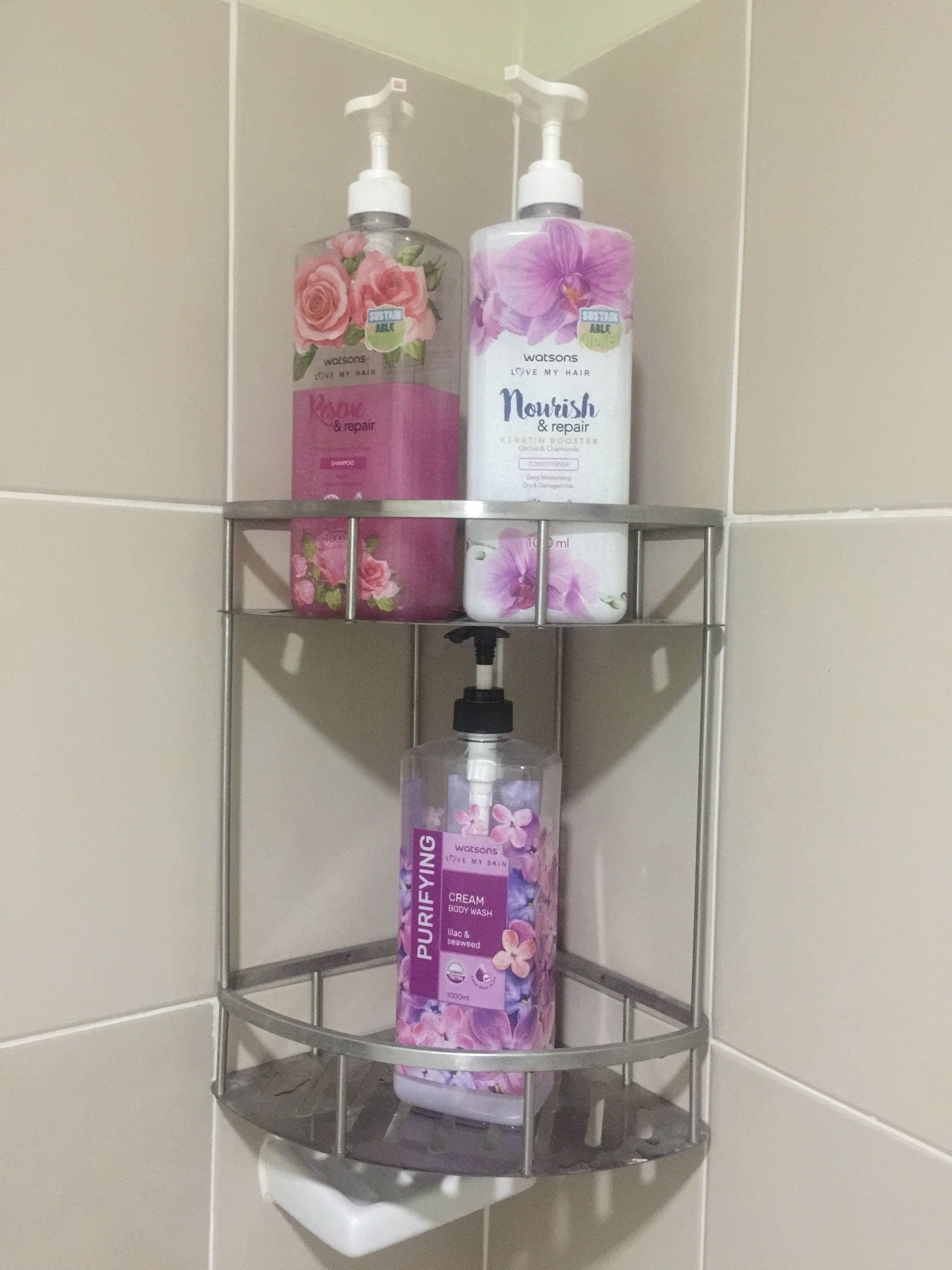 Une étagère de salle de bain avec trois flacons de produits de toilette : un shampooing rose, un après-shampooing violet et un gel douche violet.