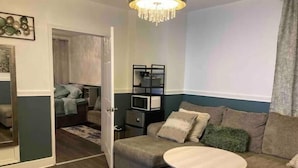 A modern living room with a sofa, a round coffee table, and a side table. There is a door leading to another room with a bed and a small table.