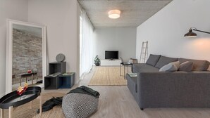Una sala moderna con un sofá gris, un televisor y una computadora portátil sobre una consola blanca. Se aprecia una pared de ladrillo y un amplio ventanal con cortinas de gasa.