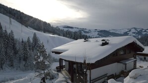 Un chalet in legno con tetto imbiancato dalla neve, immerso in un paesaggio innevato con una montagna boscosa sullo sfondo.