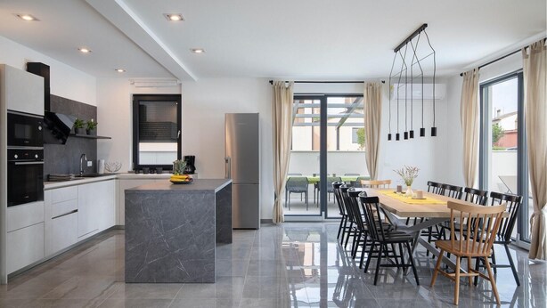 A modern kitchen with a central island, a dining area with a table and chairs, and a view of the outdoors through large windows.