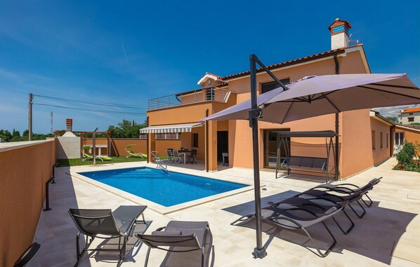 A modern outdoor pool area with lounge chairs, a covered seating area, and a two-story building with a balcony.
