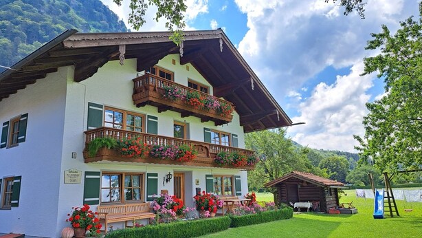Un edificio a due piani con un balcone decorato da fiori, una panchina di legno e una piccola struttura in legno nel cortile.
