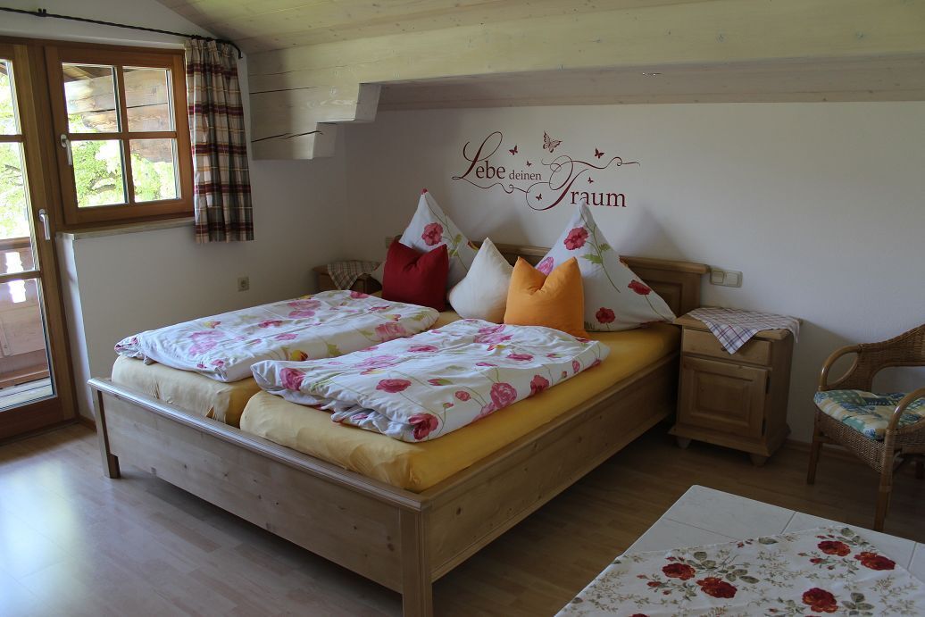A bedroom with a wooden bed, floral bedding, a bedside table, a chair, and a wall decoration.