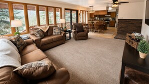 Ein geräumiges Wohnzimmer mit einem großen, braunen Ledersofa, einem Essbereich mit Holzsesseln, einer Küche mit modernen Geräten und großen Fenstern mit Blick ins Freie.