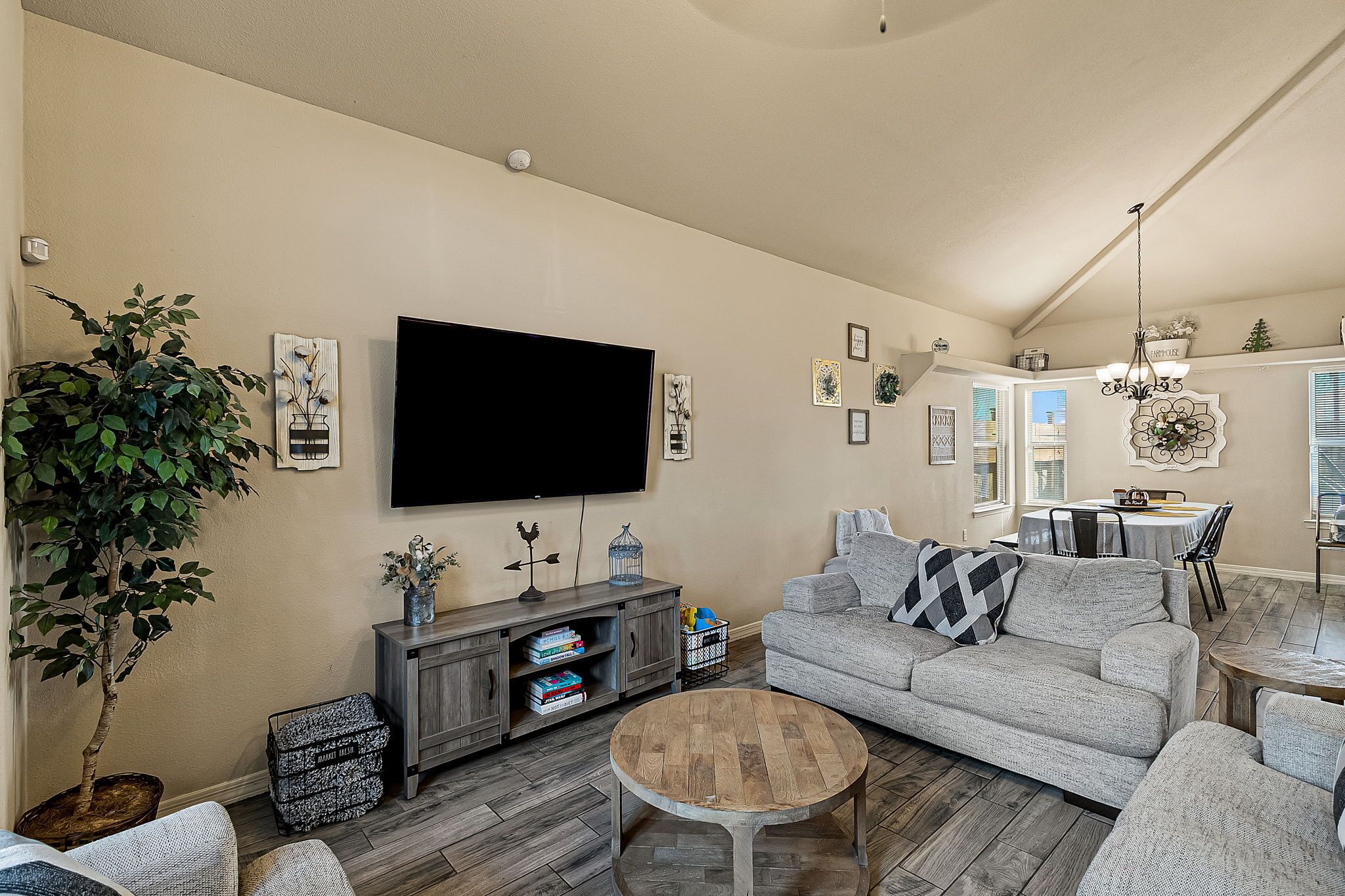 A living room with a flat-screen TV, a gray sofa, a coffee table, and a dining area with a chandelier.