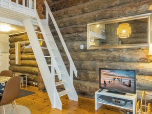 A log cabin interior with a wooden staircase, a television on a stand, and a dining area with chairs.