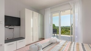 A bedroom with a bed, a TV, a wardrobe, and a balcony with a view of greenery.